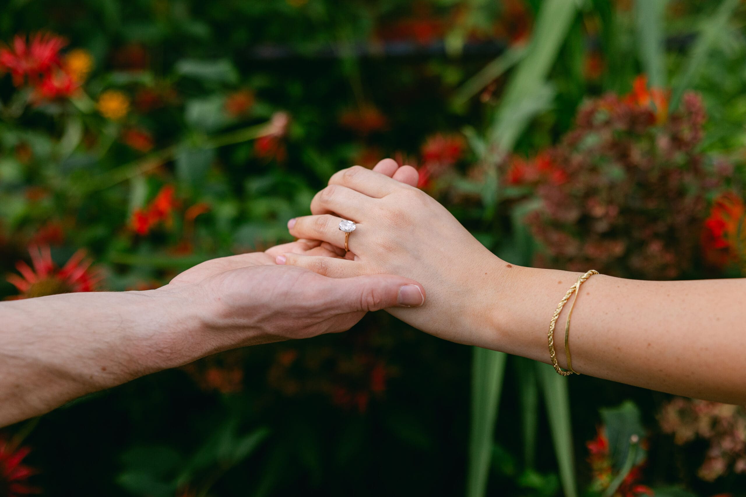 Close-up of an engaged couple holding hands, featuring an oval diamond ring against a floral backdrop