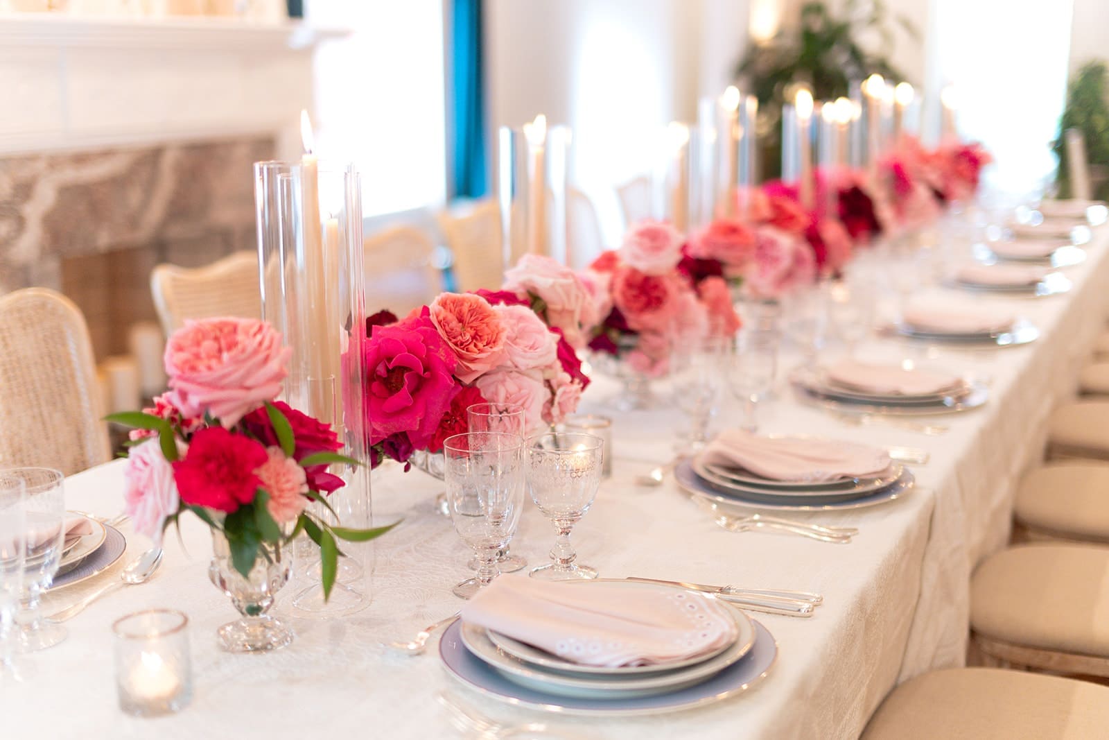 Elegant indoor wedding table lined with bright pink and berry roses and tall taper candles in glass hurricanes, creating a romantic candlelit centerpiece.
