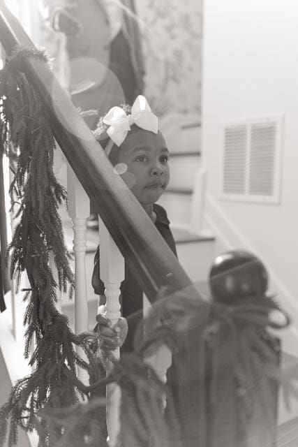 A quiet black-and-white moment of my daughter standing on the staircase beside our Christmas greenery, captured with soft light and reflective stillness.