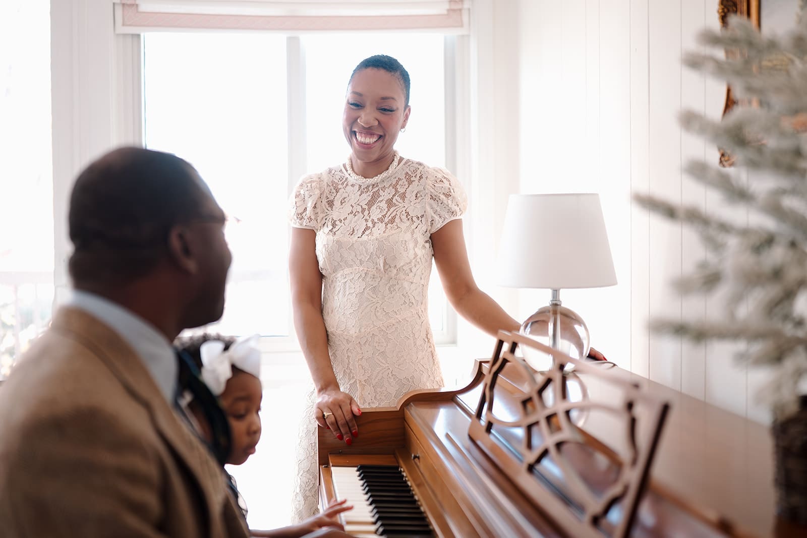 Chelsea standing beside the piano, smiling warmly as her husband and daughter play together in the morning light.