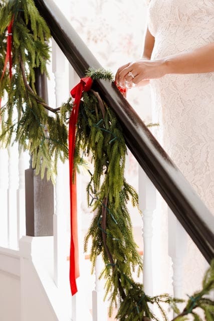 Chelsea’s hand adjusting fresh greenery and red ribbon on the staircase railing, preparing our home for the Advent season.