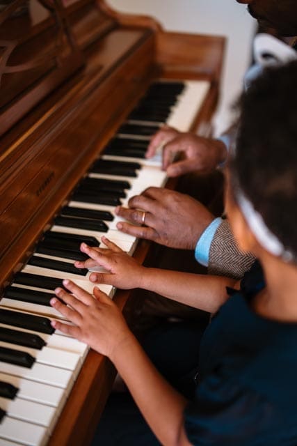Father and daughter playing the piano together, their hands side by side on the keys in a tender, everyday moment of connection.