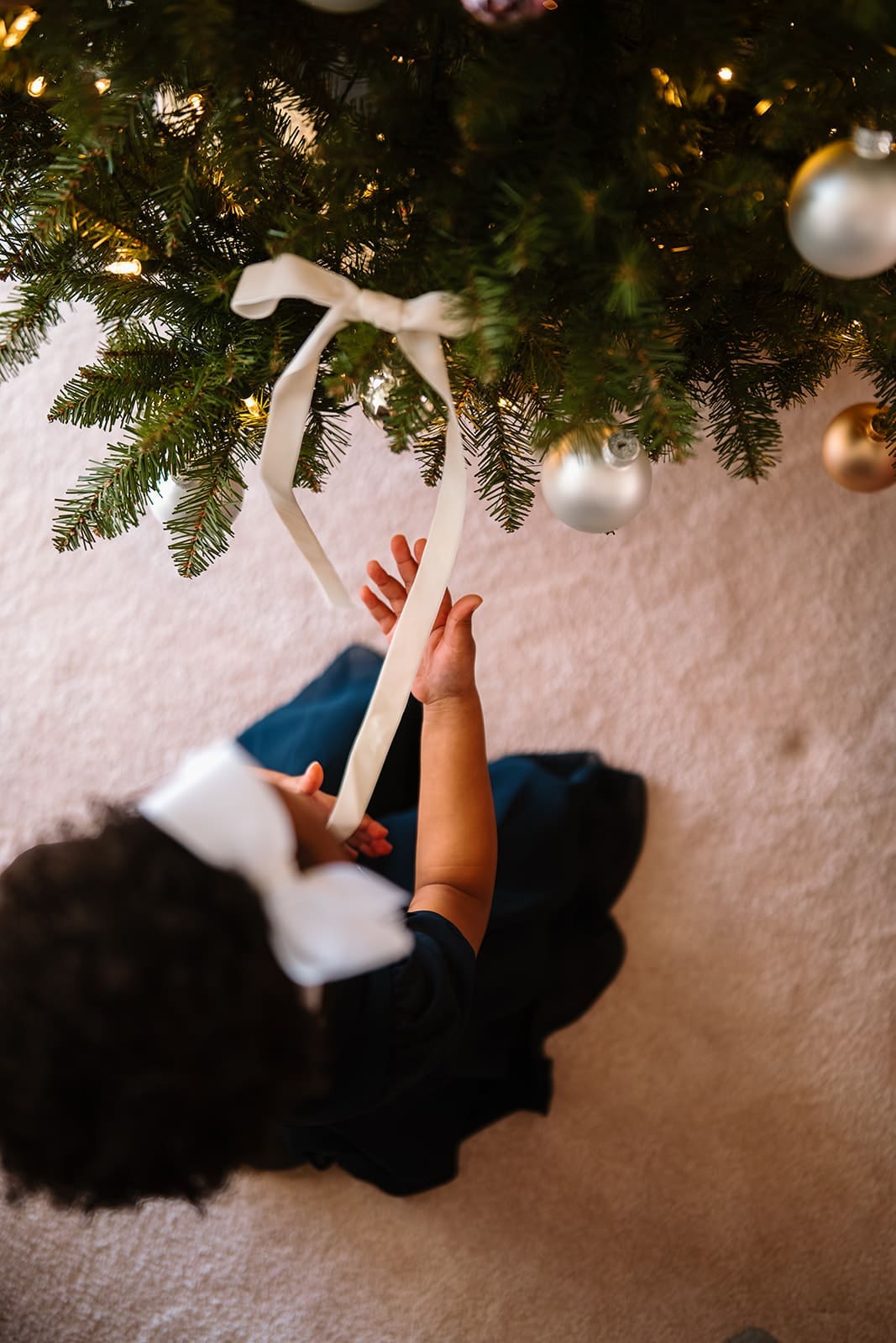 Top-down view of a young girl with a white bow holding a long ribbon beneath the Christmas tree.