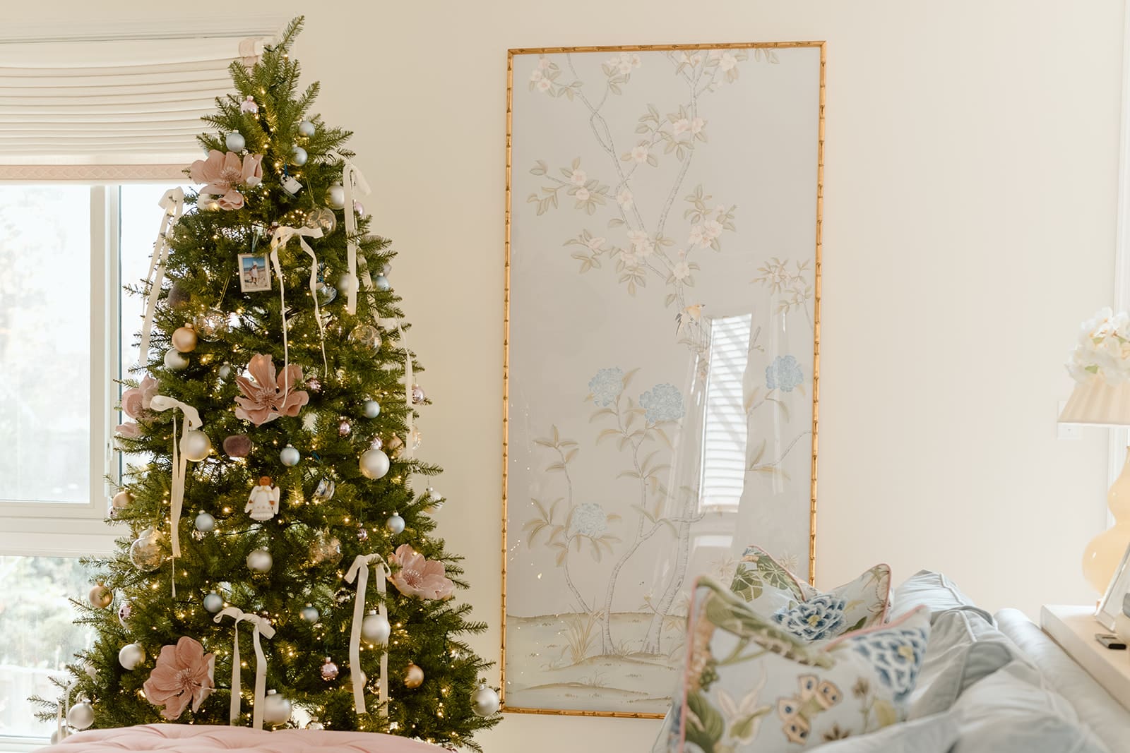 A Christmas tree decorated in soft pink, blue, and silver ornaments beside a chinoiserie wall panel and a pastel living room with a pink tufted ottoman.