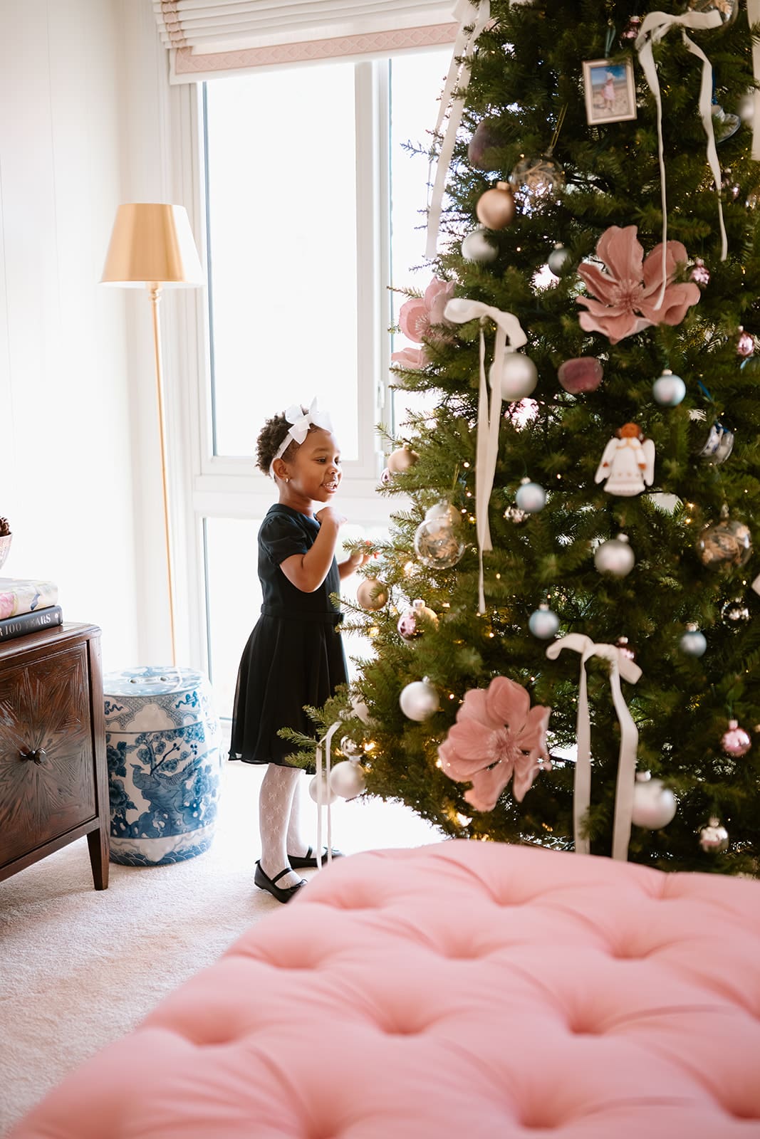 A young girl in a navy dress with a white bow gazes up at a Christmas tree decorated in soft pink and blue ornaments.