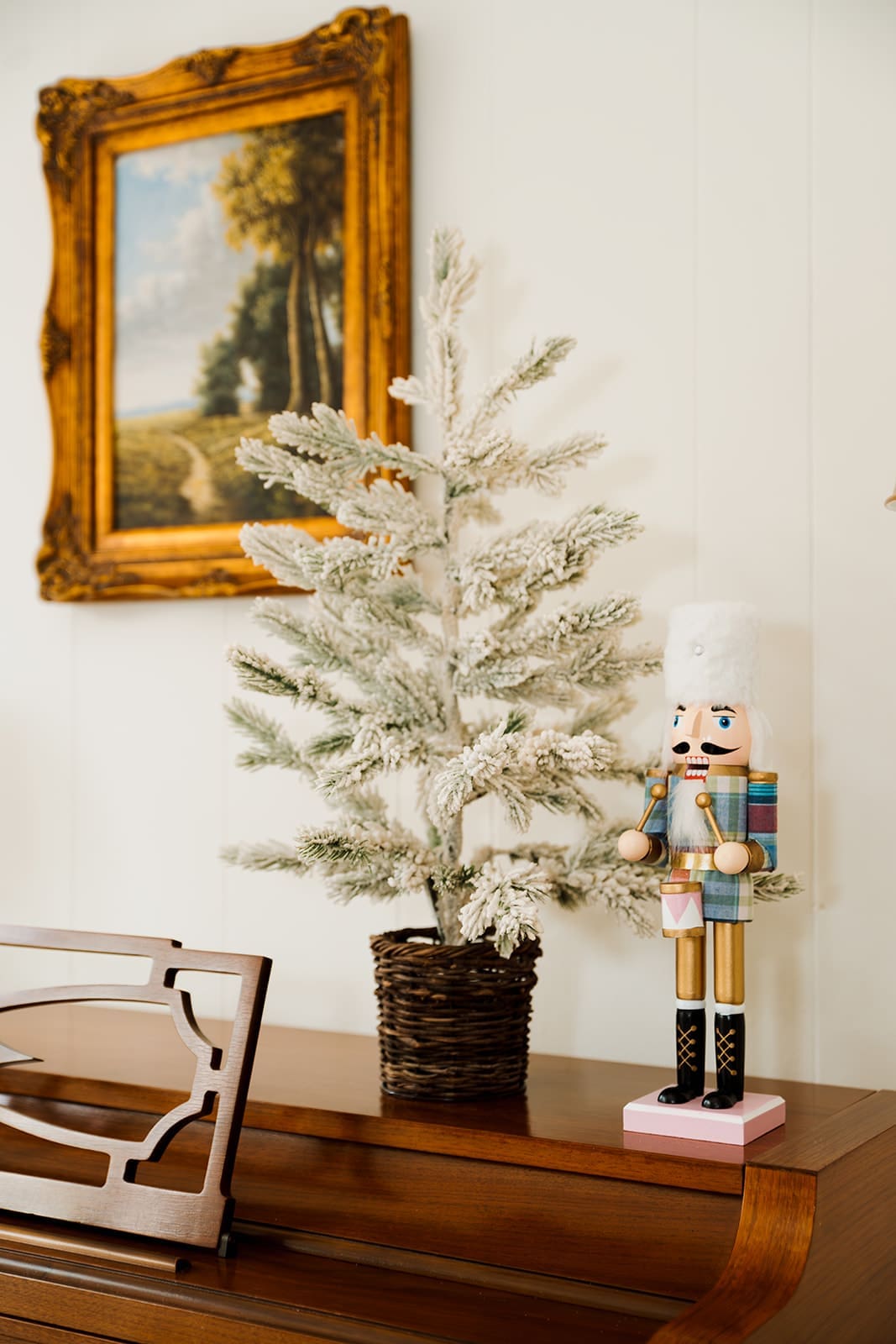 A small flocked tabletop Christmas tree and pastel nutcracker displayed on the piano, with a classic landscape painting hanging above.