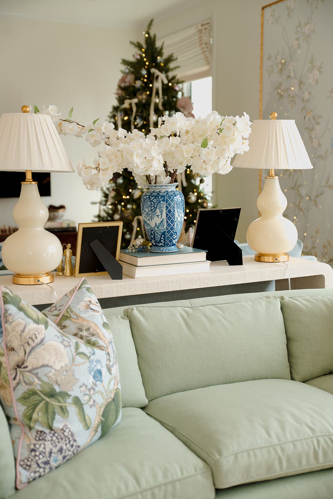 A styled console table featuring white lamps, a blue chinoiserie vase with winter florals, framed family photos, and a Christmas tree glowing in the background.