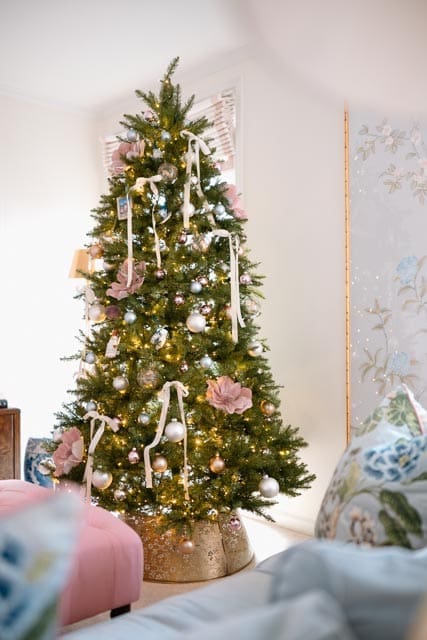A Christmas tree decorated in soft pink, blue, and silver ornaments beside a chinoiserie wall panel and a pastel living room with a pink tufted ottoman.