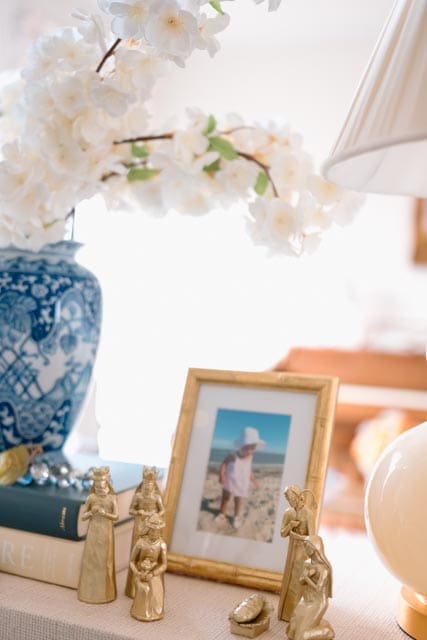 A gold nativity set styled beside a framed family photo and white floral branches in a blue-and-white vase.