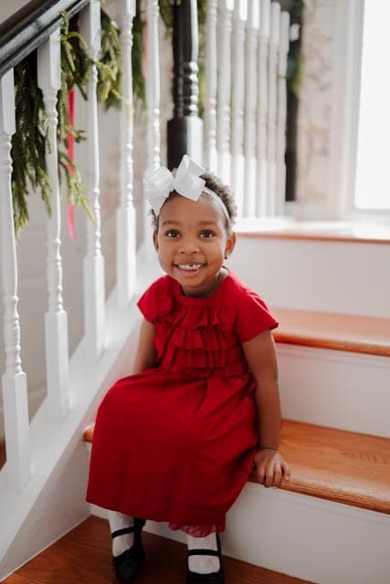 A young girl in a red dress and white bow smiles while sitting on a staircase decorated with garland and red ribbon.
