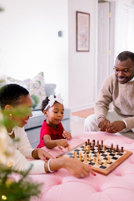 A family gathered around a chessboard on a pink tufted ottoman, smiling and playing together beside the Christmas tree.