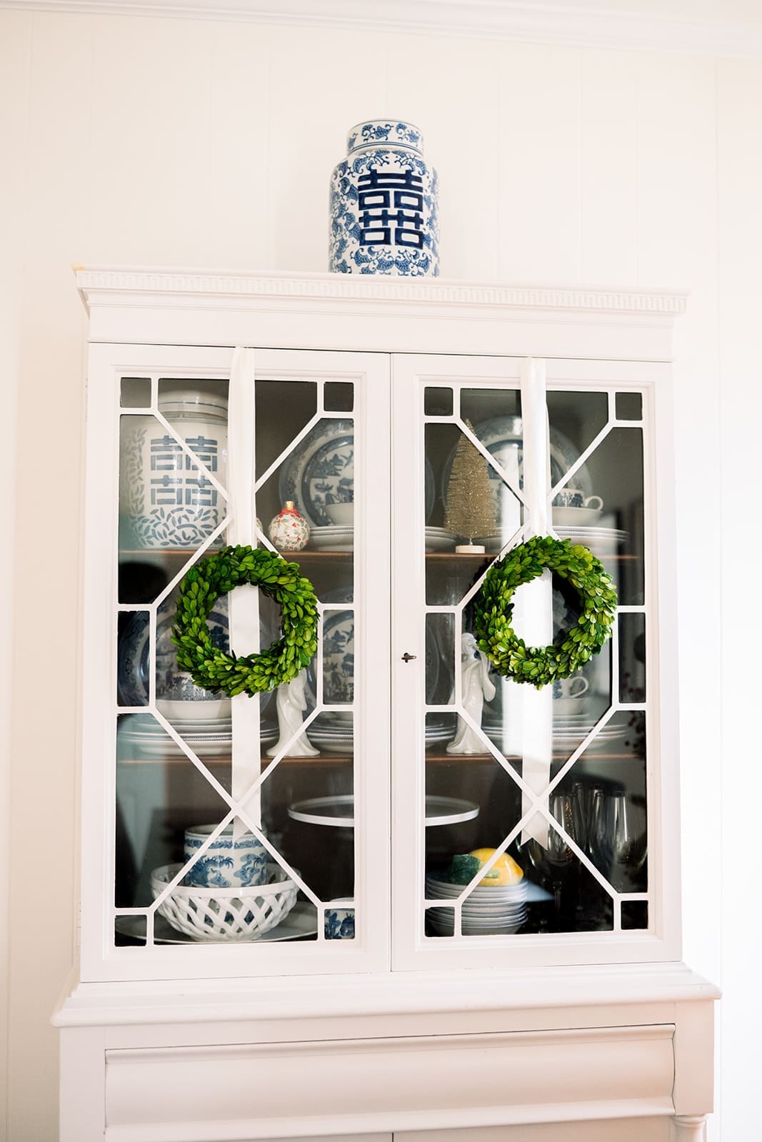 A white china cabinet decorated with two small boxwood wreaths and topped with a blue-and-white ginger jar for Christmas.
