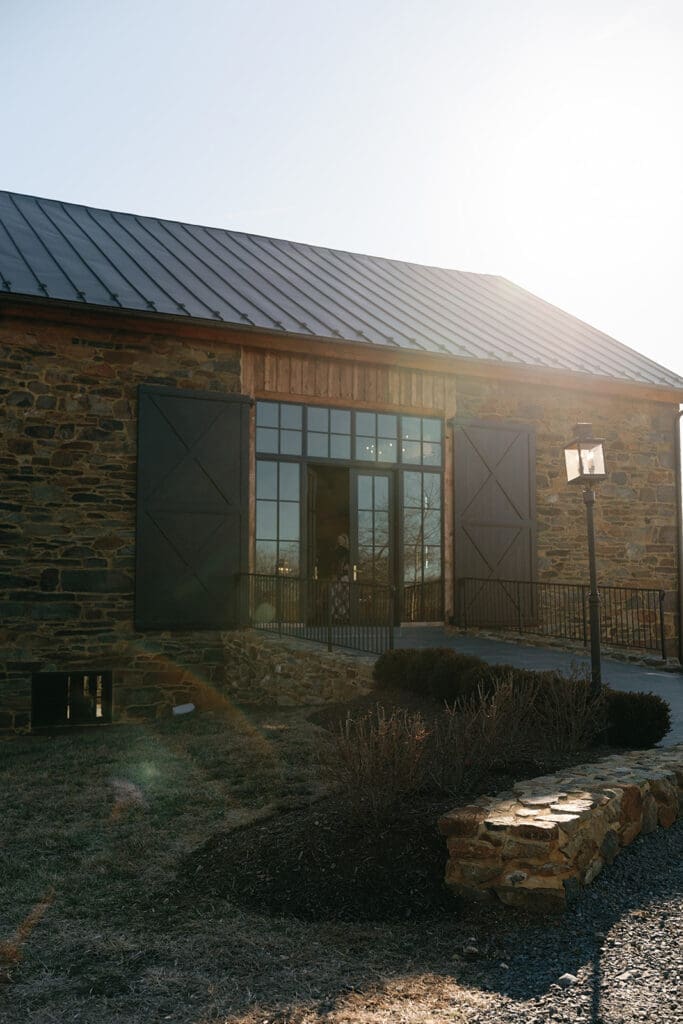 Exterior of Manor 1858 in Loudoun County with stone walls, black shutters, and warm sunlight across the entrance.