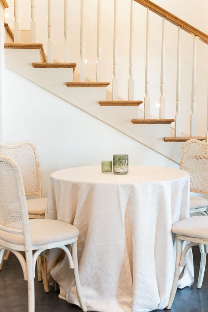 Round table with a textured ivory linen and cane-back chairs beneath the bright staircase inside The Manor 1858.
