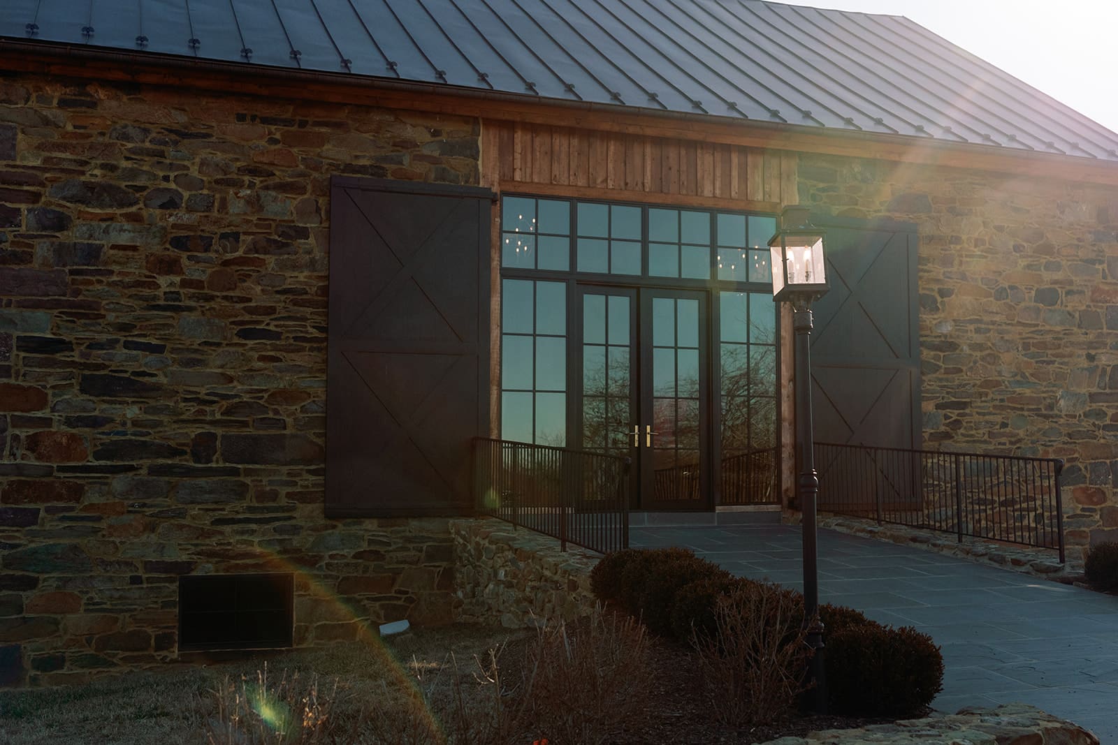 Sunlit exterior of Manor 1858 featuring the stone façade, black-framed doors, and a lantern-lined entry path.