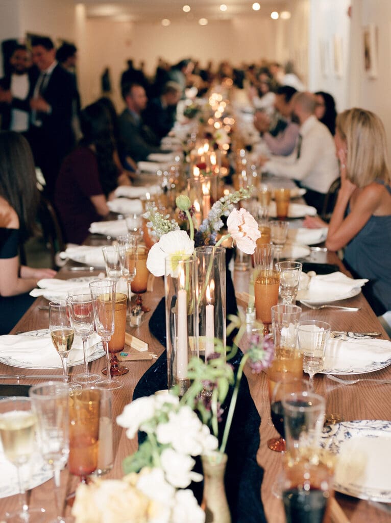 Long reception table at Fathom Gallery Georgetown set with layered glassware, candlelight, floral bud vases, and guests seated for dinner.
