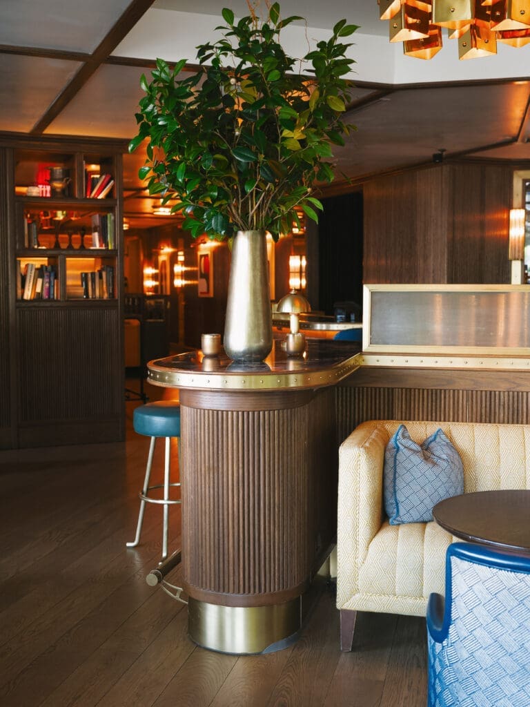 Moody bar lounge interior with dark wood paneling, brass details, blue seating, and a tall greenery arrangement