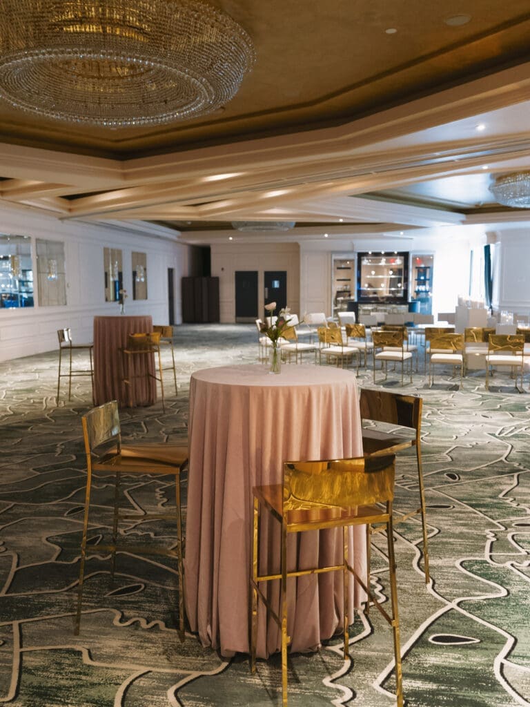 Pink cocktail table with gold bar stools in an elegant wedding reception space with ceremony setup in the background
