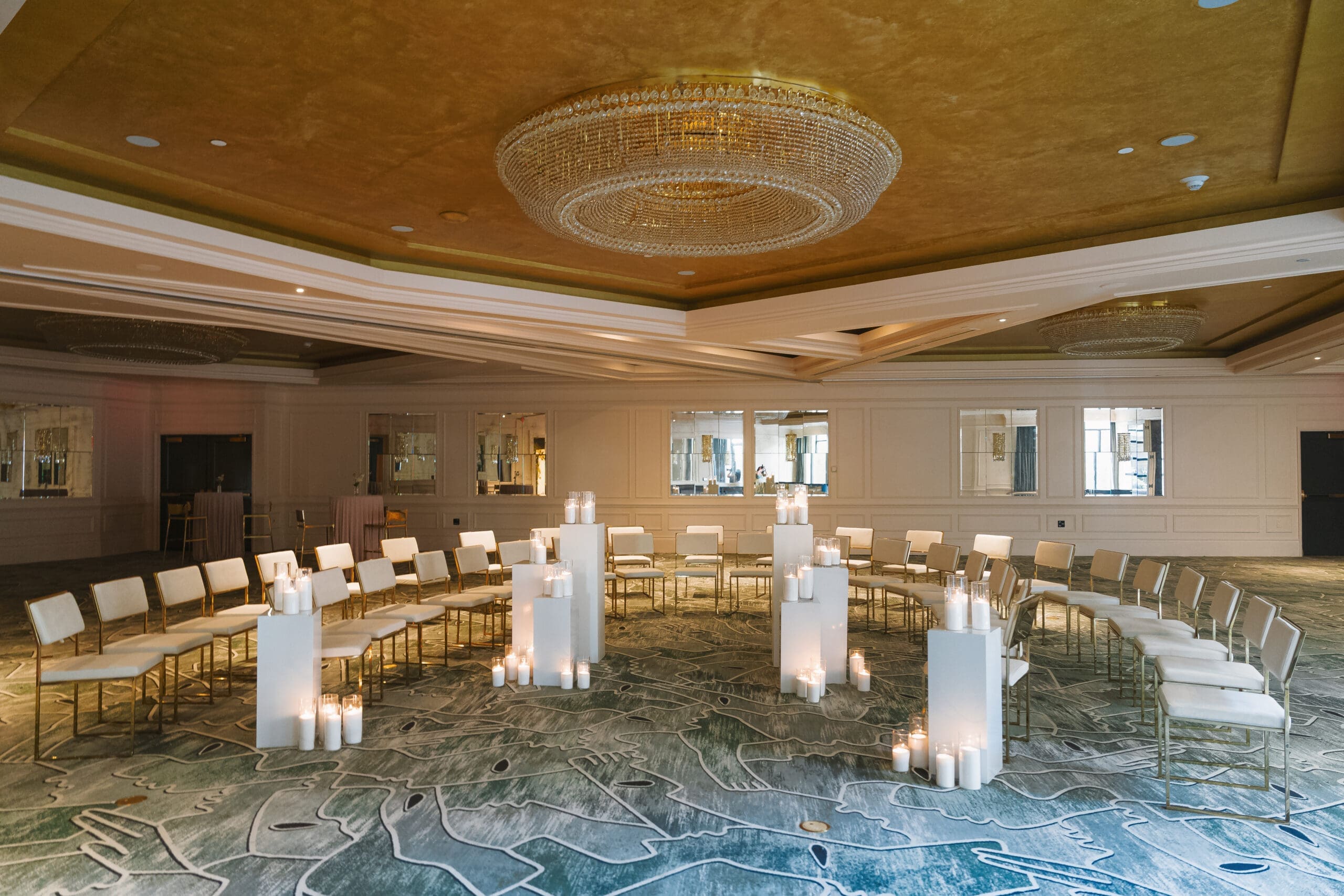 Modern indoor wedding ceremony setup with white chairs, candlelit pedestals, and a gold ceiling in an elegant ballroom